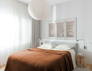 A modern minimalist bedroom featuring a bed with a brown blanket, white pillows, and a white headboard, with a large white pe