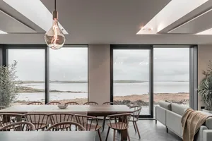 A modern residential dining room featuring a long dining table with wooden chairs, large glass windows with a coastal view, a