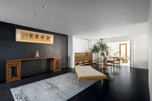A minimalist living room featuring wooden furniture, a white tufted ottoman, a black floor, and a gray patterned rug.