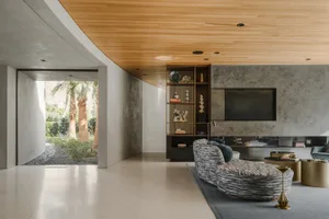 A modern living room with concrete walls, a white floor, a wooden ceiling, and a view of palm trees.