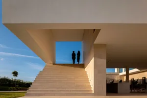 Two individuals standing on a modern, minimalist interior with stone stairs leading up to a windowed space.
