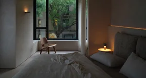 A minimally decorated bedroom featuring a textured fabric bedspread, a wooden chair, and a small table lamp with an orange li