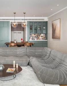 A modern open-plan living room featuring a comfortable grey upholstered L-shaped sofa, a round wooden coffee table, and a kit