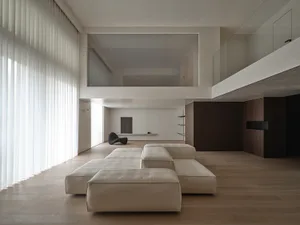 A modern minimalist living room with large windows and a wooden floor, featuring a beige modular sofa and a glass partition.