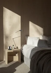 A minimalist bedroom featuring a bed with white bedding, a wooden nightstand with a lamp, and wooden walls.
