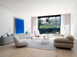 A minimalist living room with modern furniture, a large blue artwork, and a view of the outdoors through large windows.
