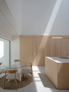 A modern minimalist residential kitchen with wood cabinetry, a glass dining table, and a large window allowing natural light.