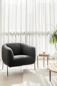 A modern living room with a comfortable fabric chair, white sheer curtains, and a small wooden table with a candle and book.