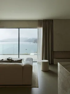 Modern minimalist living room with a large window overlooking the water, featuring a beige fabric sofa and a glass coffee tab