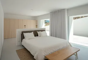 A minimalist bedroom with a large bed, white curtains, and wooden furniture.