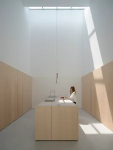 Person preparing food in a minimalist modern kitchen with wood cabinets, white countertops, and overhead lighting.