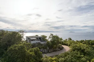 Modern architectural home with concrete walls and a glass terrace overlooking the ocean from a rocky cliffside.