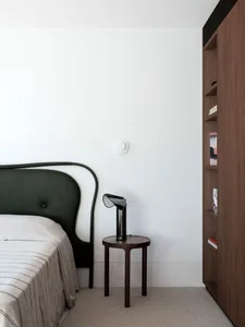 A minimalist bedroom featuring a black headboard bed, a wooden bedside table with a black lamp, and a wooden bookshelf.