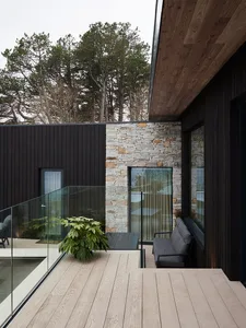 Modern outdoor living space featuring a wooden deck with a glass railing and a stone wall, surrounded by trees.