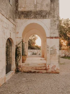 A stone entryway with an arched opening, leading to a residential courtyard with plants and trees in the background.