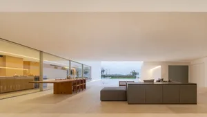A spacious, minimalist residential interior featuring light-colored wood, glass walls, and concrete elements.