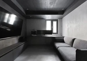 An industrial-style living room with concrete walls and floor, featuring a grey sofa and a large TV on the left wall.