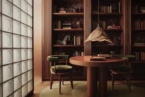 A modern library study space with wooden shelves, round table, and green velvet chairs.