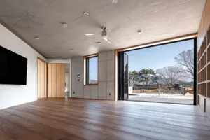 A modern minimalist living room with concrete walls, large windows, and a wooden floor.