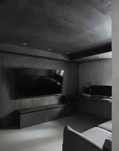 Modern industrial-style living room with concrete walls and floor, featuring a flat-screen TV and grey upholstered furniture.