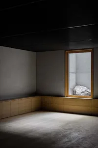 A minimalist bedroom with concrete walls and flooring, featuring a large window with a rock installation.