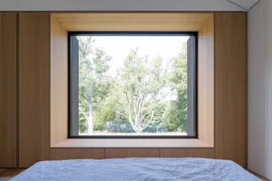 A minimalist bedroom with a large window framed in wood, overlooking a forested area.
