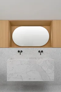 A minimalist bathroom with white marble floating vanity, black faucets, white hexagonal tiles, and light wood cabinets.