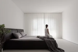 A minimalist bedroom with black furniture, grey sheets, and a woman sitting on the bed looking out through vertical blinds.