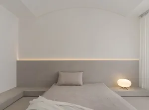 Minimalist bedroom with concrete walls and floor, featuring a bed with simple bedding and a small round lamp.