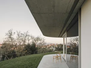A minimalist house with a concrete exterior features a sleek, angular roof and large glass window overlooking a serene landsc