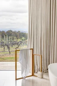 A spacious modern bathroom with long beige curtains, a towel rack, a bathtub, and a view of a vineyard.