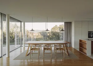 A modern living-dining room featuring a wooden dining table and chairs, with a large glass window showcasing a scenic outdoor