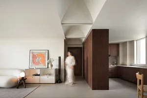 A minimalist modern living room featuring a white sofa, a wooden pillar, and a kitchen area with dark wooden cabinets.