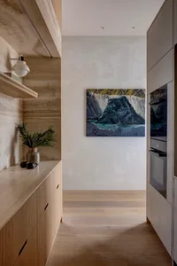 A modern minimalist kitchen with concrete walls and wood cabinets, featuring a painting of a mountain landscape.