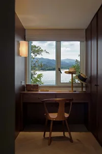 Modern residential space featuring windows overlooking a serene lake view, with wooden furniture and a wooden table lamp.