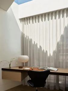 A minimalist home office featuring a wooden desk, black chair, open book, and decorative vase with flowers under a large curt