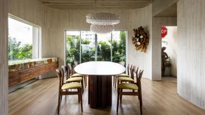 A modern dining room with a wooden table, glass chandelier, and greenery outside.
