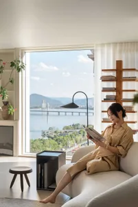 A woman sitting on a beige couch reading a book in a modern neutral-toned living room with a view of a river.