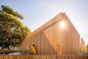 A woman in a yellow dress and hat stands outside a modern architectural building made of wood.