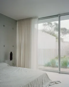 A minimalist bedroom with white walls, a bed covered in white sheets, and large windows with sheer curtains allowing natural 