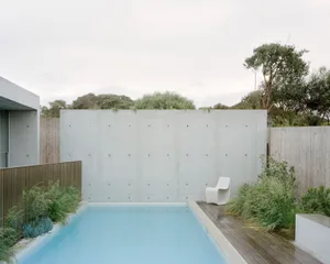 A modern minimalist pool side seating area with a white chair on a wooden deck surrounded by greenery.