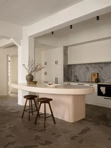 A minimalist residential kitchen featuring a concrete island with a smooth, curved countertop, two wooden stools, a vase, and