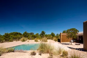 A modern house with a wooden exterior and a swimming pool surrounded by sand and vegetation.