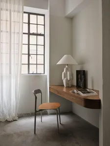 A minimalist office space featuring a wooden desk setup with a white lamp, open book, and portrait on the wall, alongside she