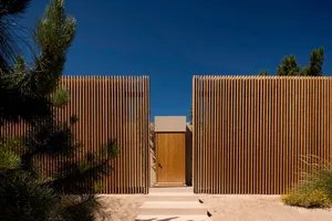 A modern wooden fence with steps leading to a door, surrounded by sandy ground and natural vegetation.