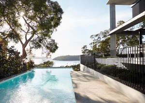 A modern outdoor residential area featuring a pool with a view of the sea, surrounded by greenery and a sleek, contemporary h