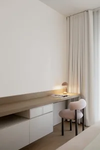 Minimalist residential room featuring a wooden desk with a lamp, a cushioned chair, and floor-to-ceiling curtains.