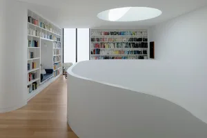 A modern library interior with white concrete walls, shelves filled with books, and an oval skylight.
