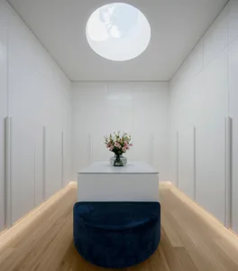 A minimalist interior space featuring white panels on the walls and ceiling, with a circular skylight, a small table with a v
