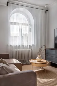 Modern minimalist living room with neutral-toned furniture and decor, soft lighting, and a large window with white sheer curt